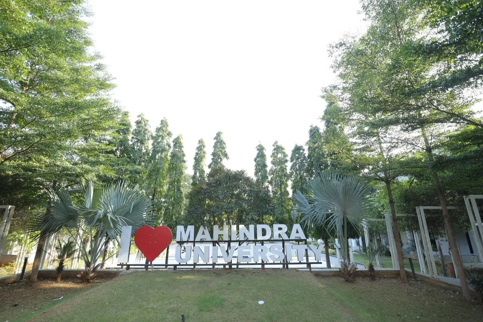 The “I ♥ Mahindra University” sign on the Mahindra University campus, framed by tall trees and palms.